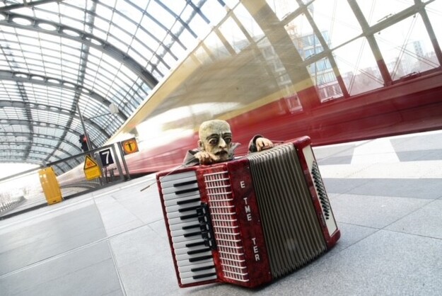 Puppe mit Akkordeon am Bahnsteig eines Bahnhofs