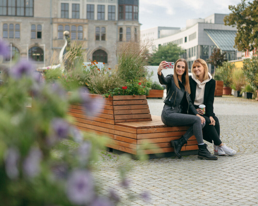 Zwei junge Frauen machen ein gemeinsames Bild in der Gartenträume Lounge in Dessau. Im Hintergrund ist das Rathauscenter und das Rathaus zu sehen