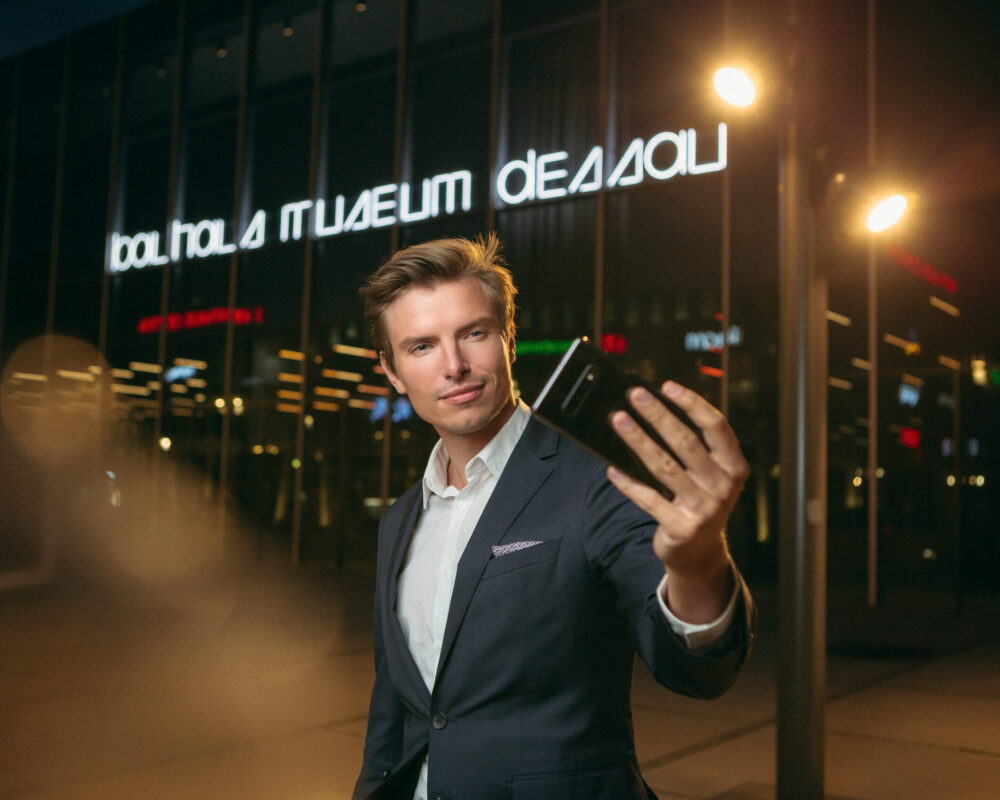 Das Bild zeigt einen Mann im Anzug, der ein Selfie vor dem beleuchteten Bauhausmuseum in Dessau macht